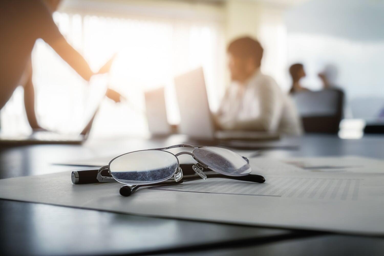 Óculos sobre documentos com equipe desfocada ao fundo em reunião de negócios