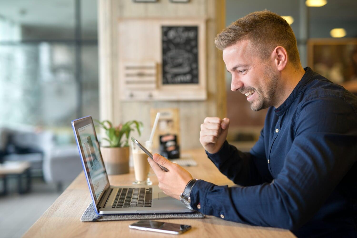 Homem comemorando ao usar um celular em frente a um notebook em ambiente de trabalho moderno